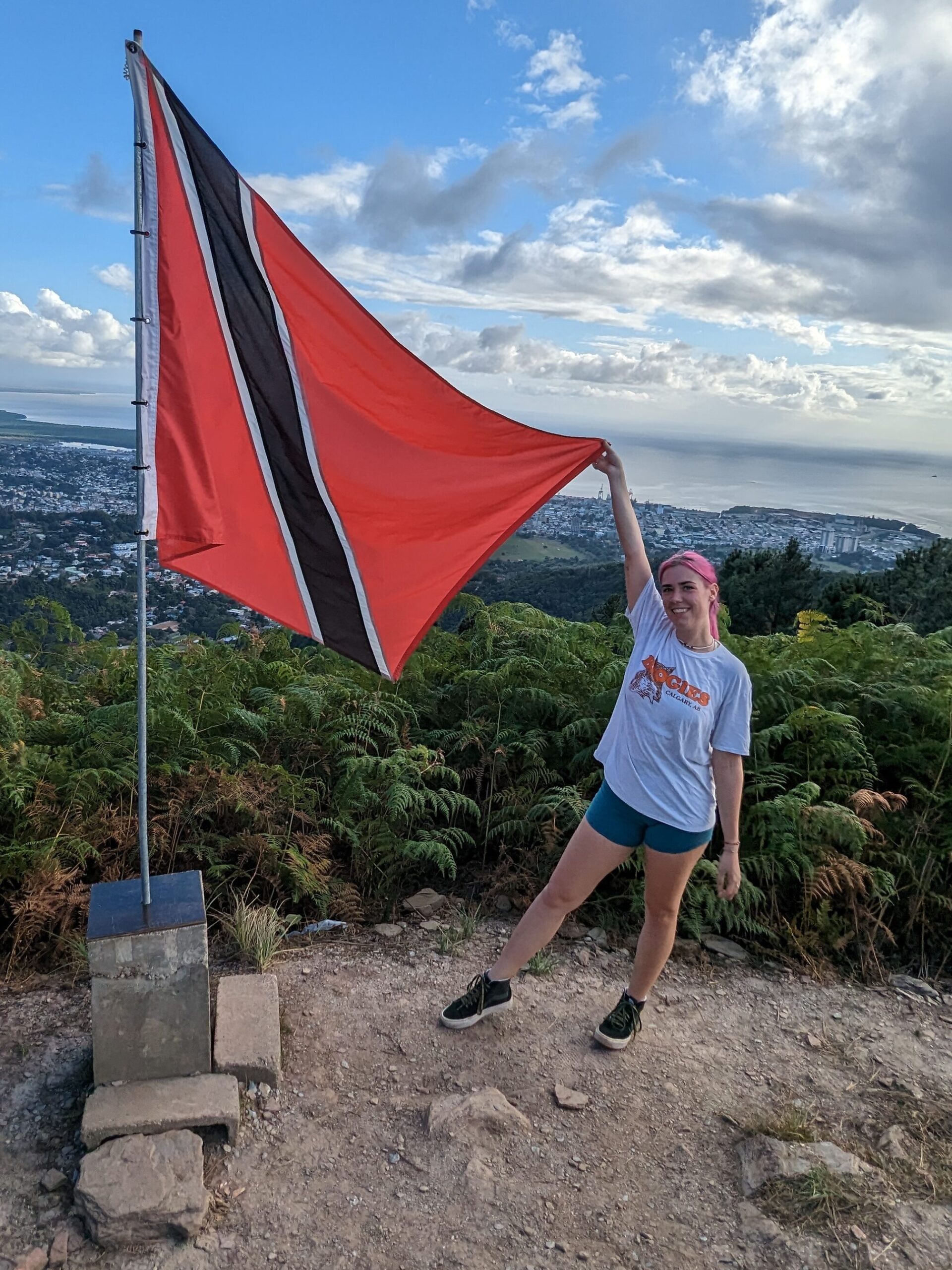 Emily stands atop a mountain, holding the flag of Trinidad and Tobago, overlooking the countryside.